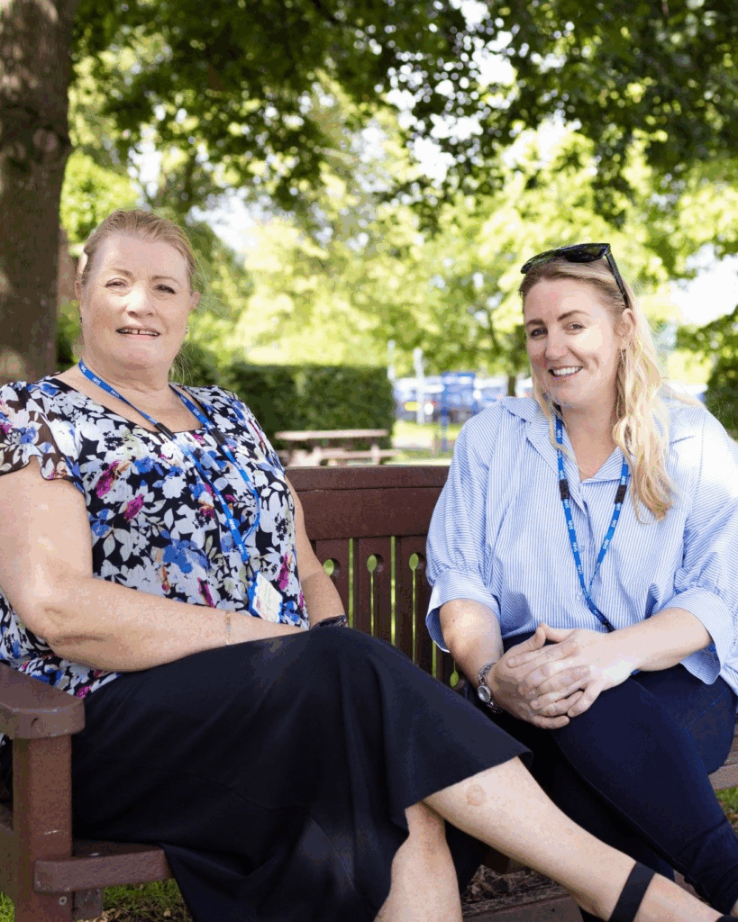Two women, both wearing blue NHS lanyards, are sitting and smiling on a wooden bench in a sunlit park. Trees and a picnic bench are visible in the background, suggesting a relaxed and pleasant outdoor environment.