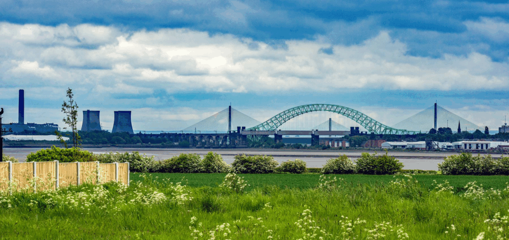 A scenic view across a river with green fields in the foreground. In the background are two prominent bridges—the Silver Jubilee Bridge (green arch) and the Mersey Gateway Bridge (cable-stayed)—alongside industrial buildings and cooling towers under a cloudy sky.