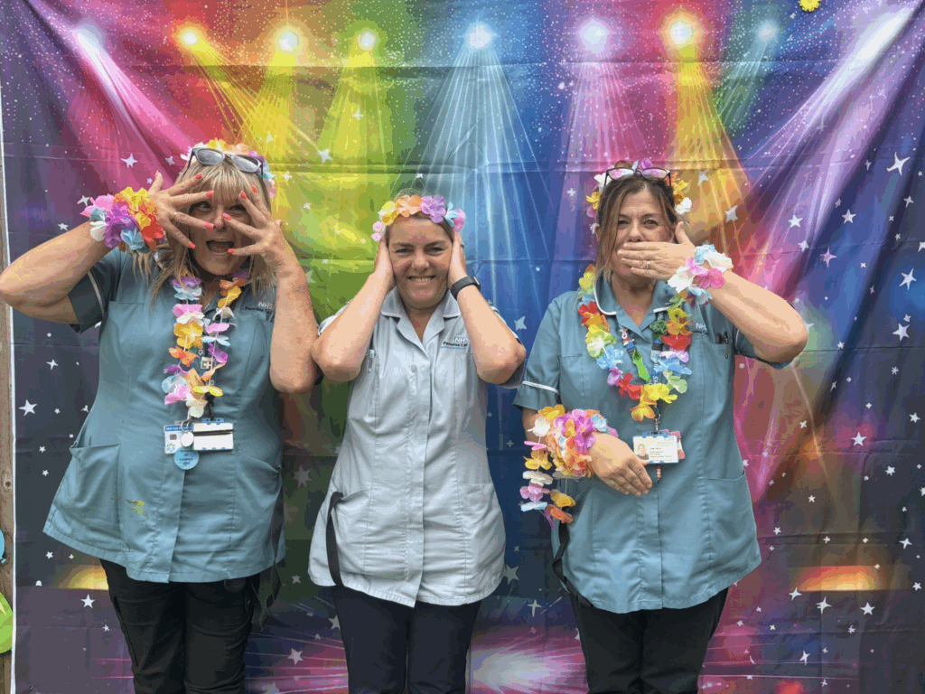 Three women dressed in NHS uniforms pose playfully in front of a colourful, backdrop with rainbow lights. They wear flower garlands and perform the "see no evil, hear no evil, speak no evil" gesture, smiling and having fun.