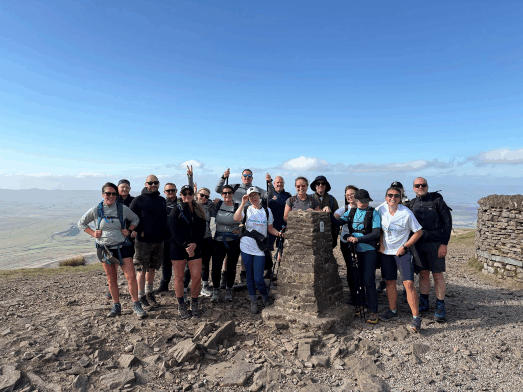 A large group of Pennine Trust hikers poses at the summit of a hill or mountain on a clear sunny day, with expansive views of the countryside in the background, after a charity sponsored mental health hike. They are gathered around a stone marker, wearing outdoor gear and smiling at the camera.