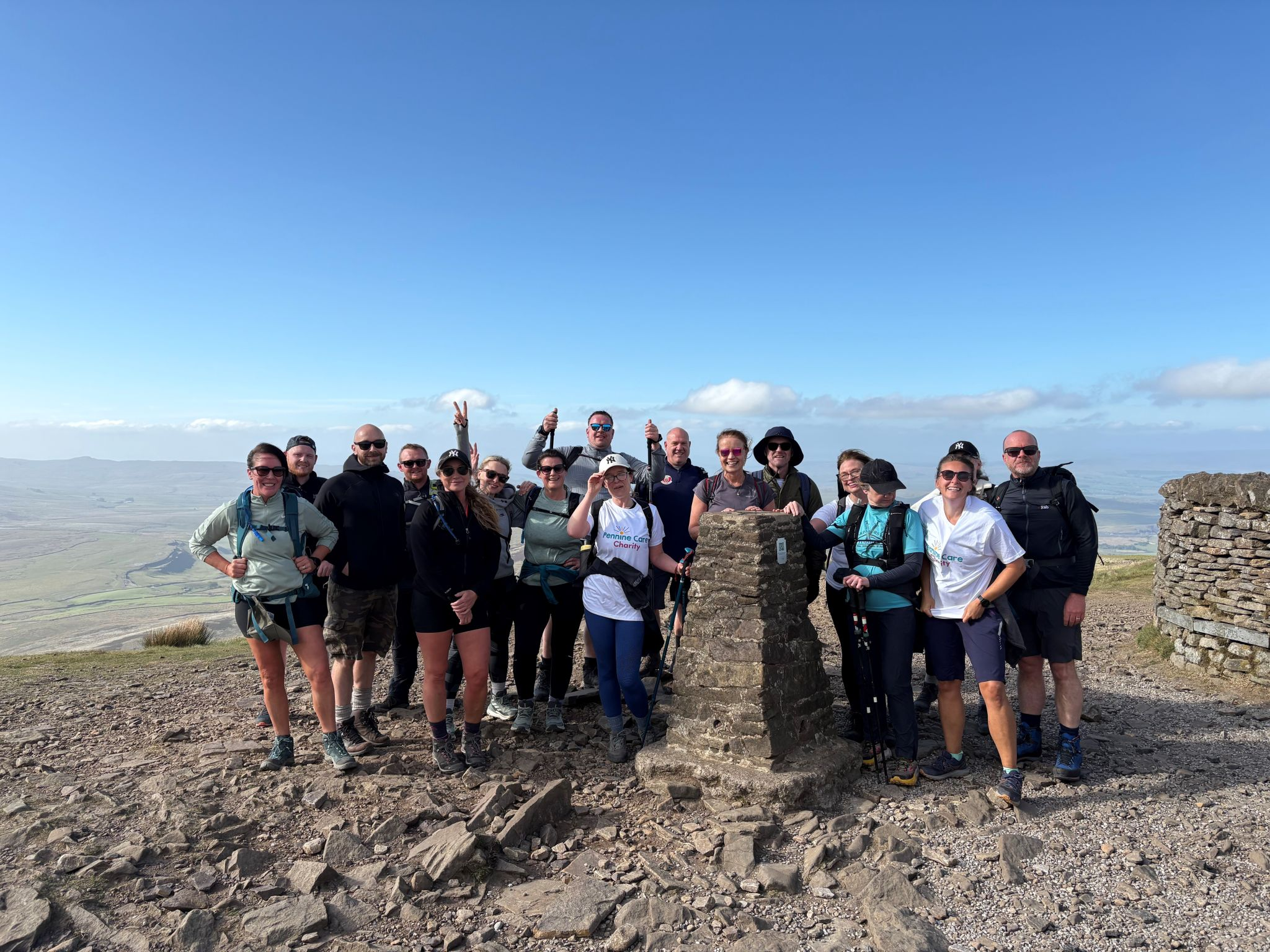 A large group of Pennine Trust hikers poses at the summit of a hill or mountain on a clear sunny day, with expansive views of the countryside in the background, after a charity sponsored mental health hike. They are gathered around a stone marker, wearing outdoor gear and smiling at the camera.