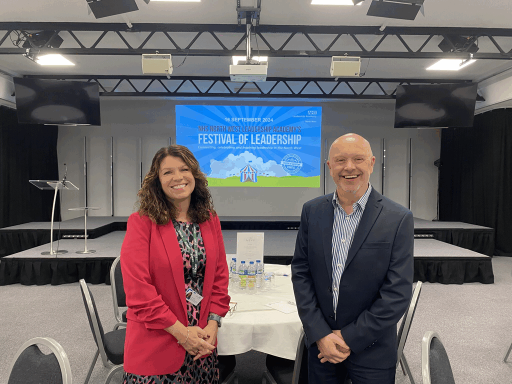 Frances Dodd, Head of the NHS North West Leadership Academy, and Chris Cutts from NHS England North West standing in front of a stage at the NHS North West Leadership Academy’s Festival of Leadership 2024 event. The event screen behind them reads ‘Connecting, celebrating and inspiring leadership in the North West.