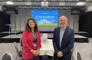 Frances Dodd, Head of the NHS North West Leadership Academy, and Chris Cutts from NHS England North West standing in front of a stage at the NHS North West Leadership Academy’s Festival of Leadership 2024 event. The event screen behind them reads ‘Connecting, celebrating and inspiring leadership in the North West.