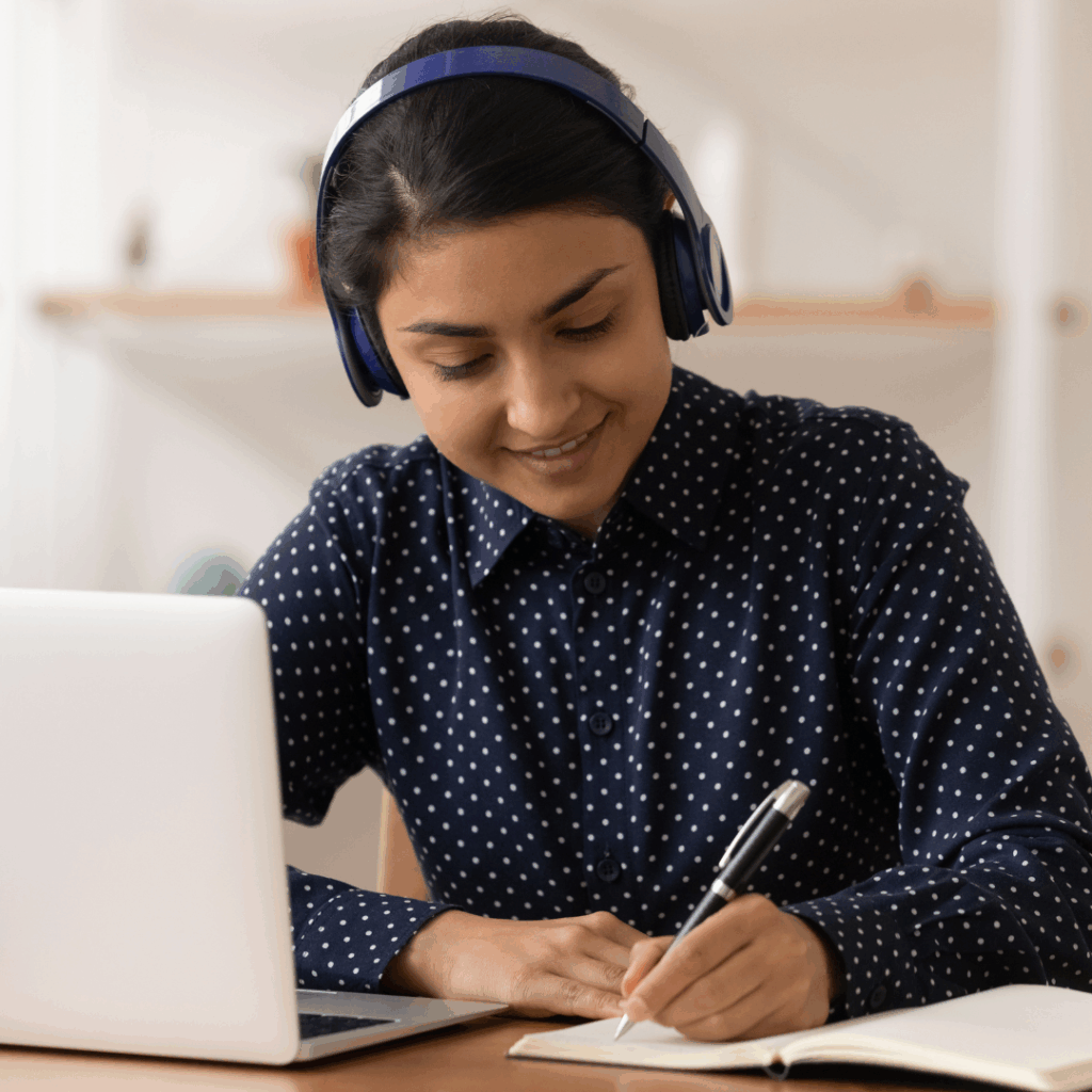 A person wearing headphones sitting at a desk with laptop writing notes.
