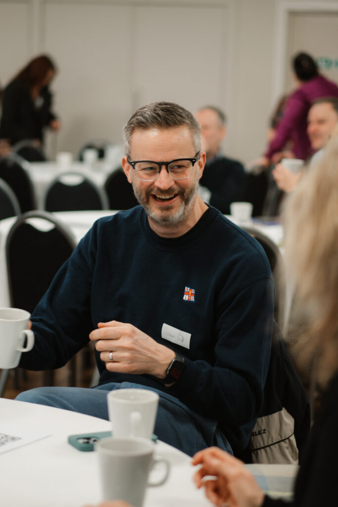 A person with a blurred face sits at a table holding a white mug, wearing a dark blue sweater with a small emblem and a name tag. In the background, other individuals with blurred faces are seated at tables covered with white tablecloths and surrounded by black chairs, suggesting an indoor event or meeting space.