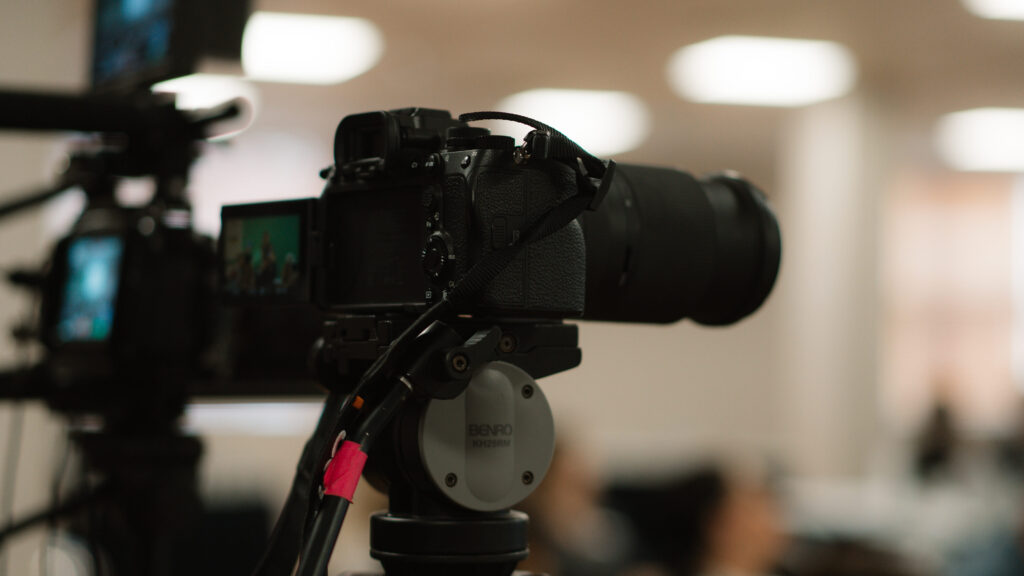 A professional camera mounted on a tripod in an indoor setting, positioned for filming or photography. The background is softly blurred, revealing several people and bright ceiling lights.