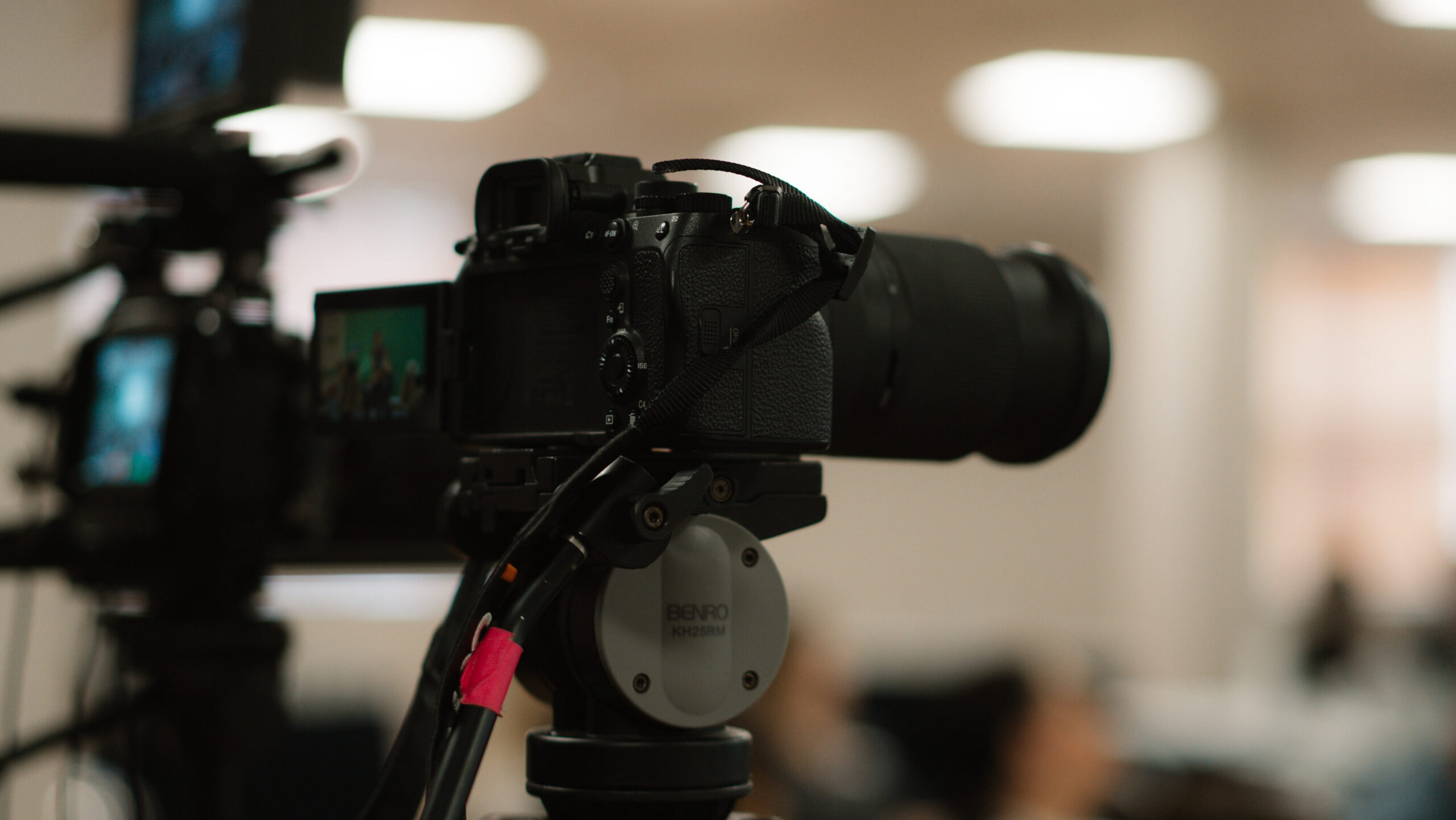 A professional camera mounted on a tripod in an indoor setting, positioned for filming or photography. The background is softly blurred, revealing several people and bright ceiling lights.