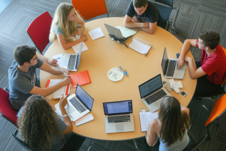 Six individuals sitting around a large round table in a collaborative workspace. Each person has a laptop open, and there are notebooks, papers, and writing materials scattered across the table. The floor has a patterned carpet, and the chairs are in various colors, including red and orange.